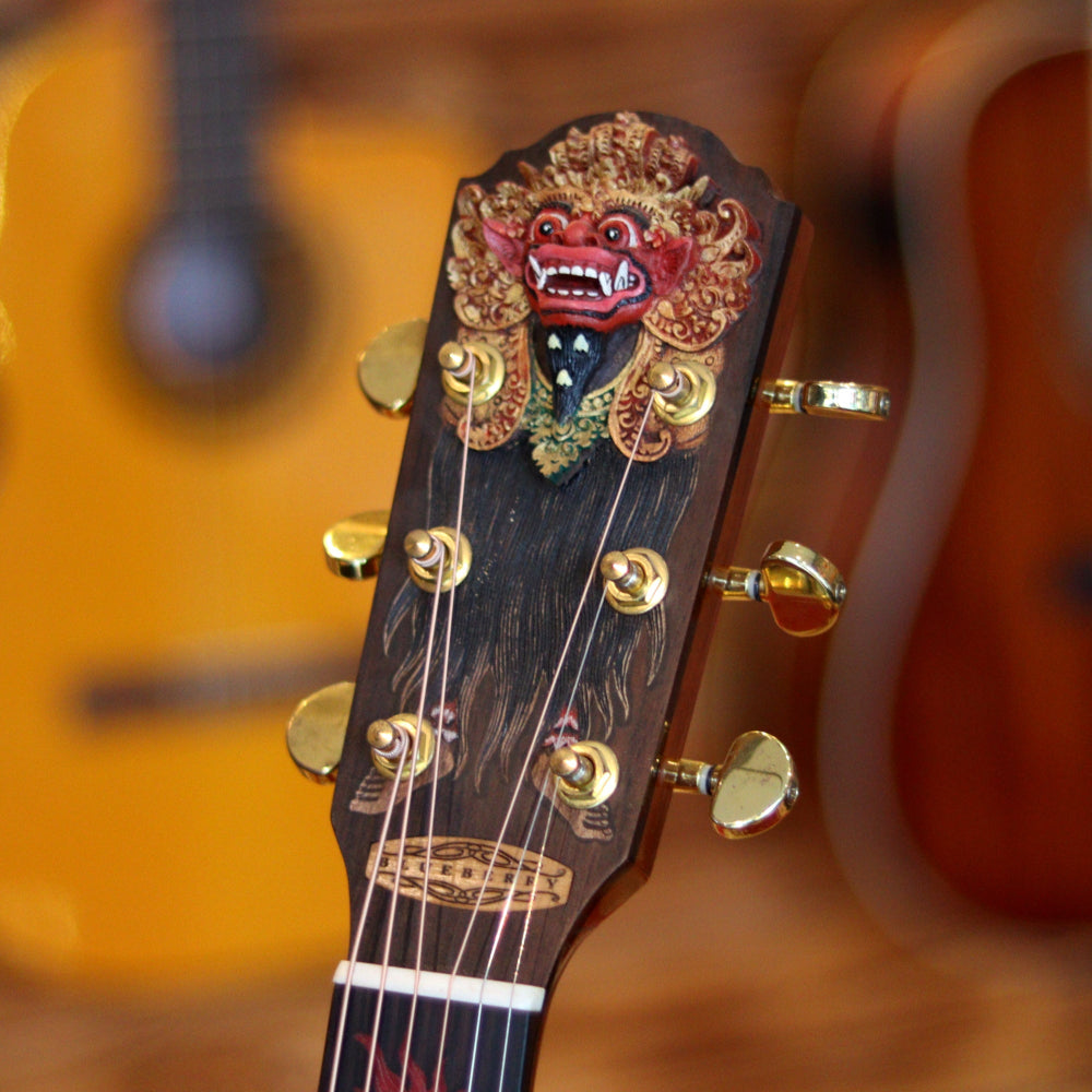 A guitar headstock with gold tuning pegs and a red Barong face decoration at the top