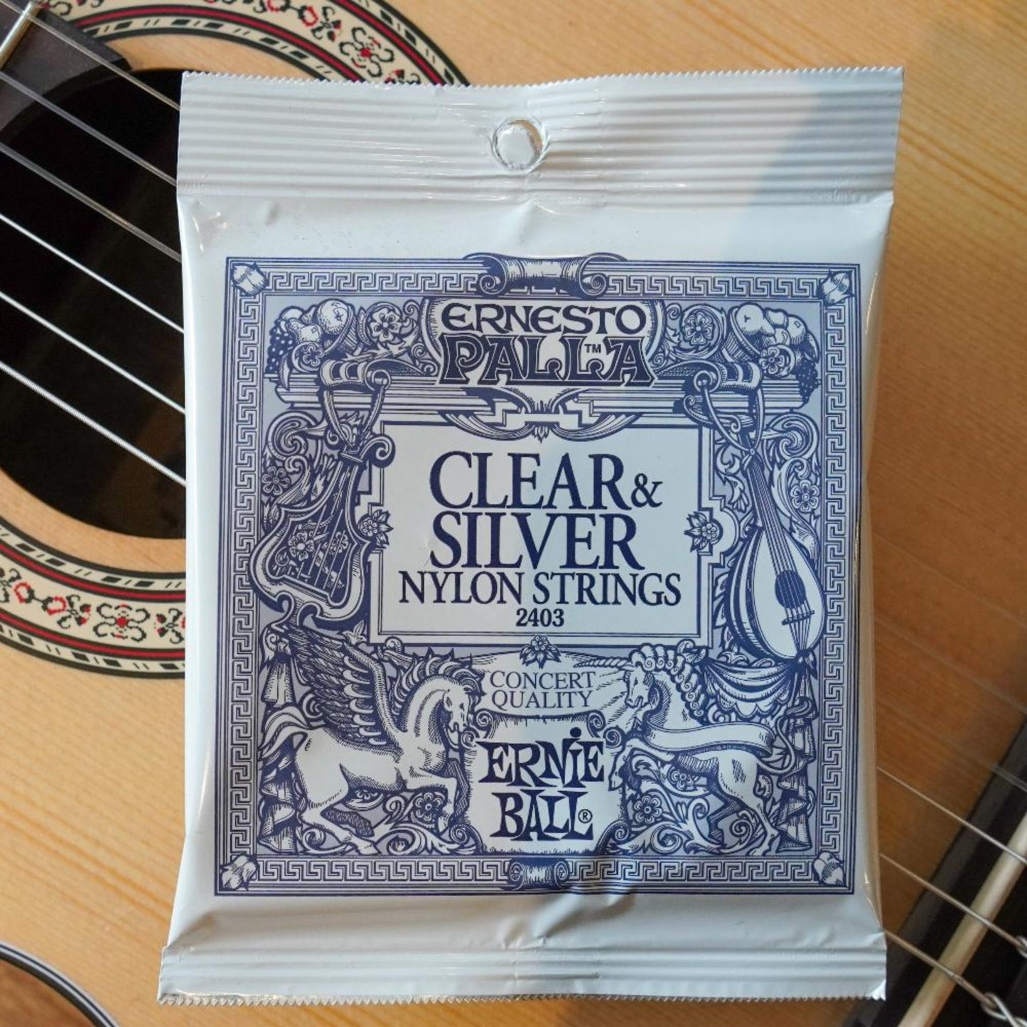 A pack of Ernie Ball Ernesto Palla clear and silver nylon strings for guitar, laid out in front of a guitar.