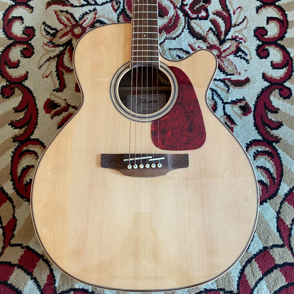 Close-up of the body of a Takamine GN93CE Natural acoustic guitar on a red carpet, showing its glossy finish, rosette, and red pickguard.