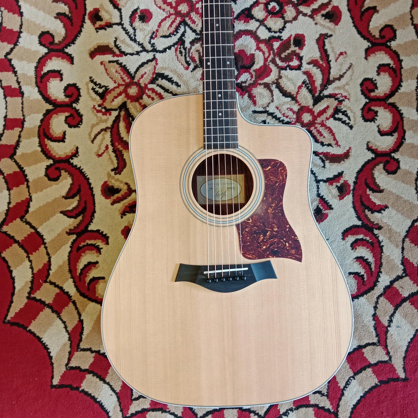 Front body of a Taylor 210CE Dreadnought acoustic guitar with a Sitka spruce top, displayed on a patterned red and beige backdrop.