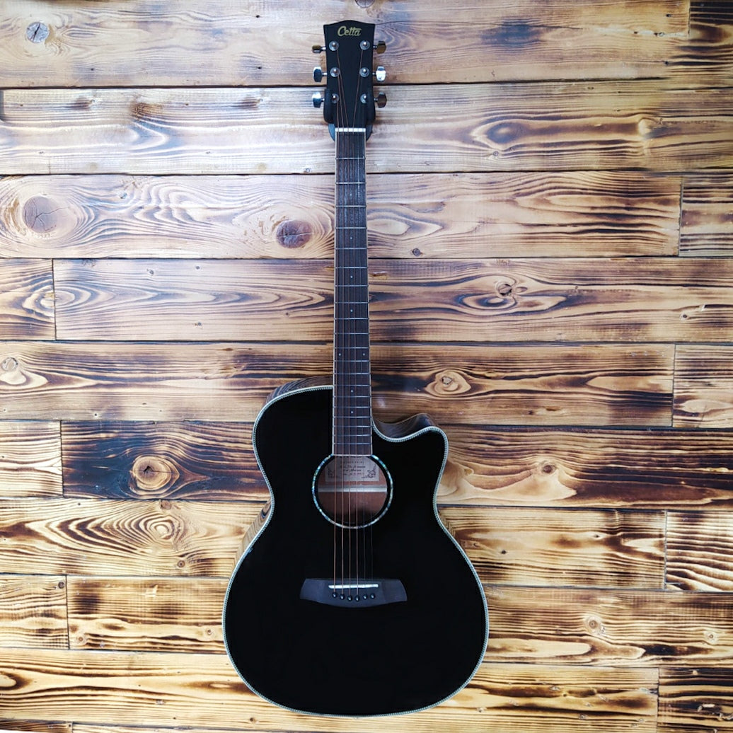 A Cetta OM28CE acoustic guitar resting on a wood wall, featuring a natural wood finish and cutaway design.