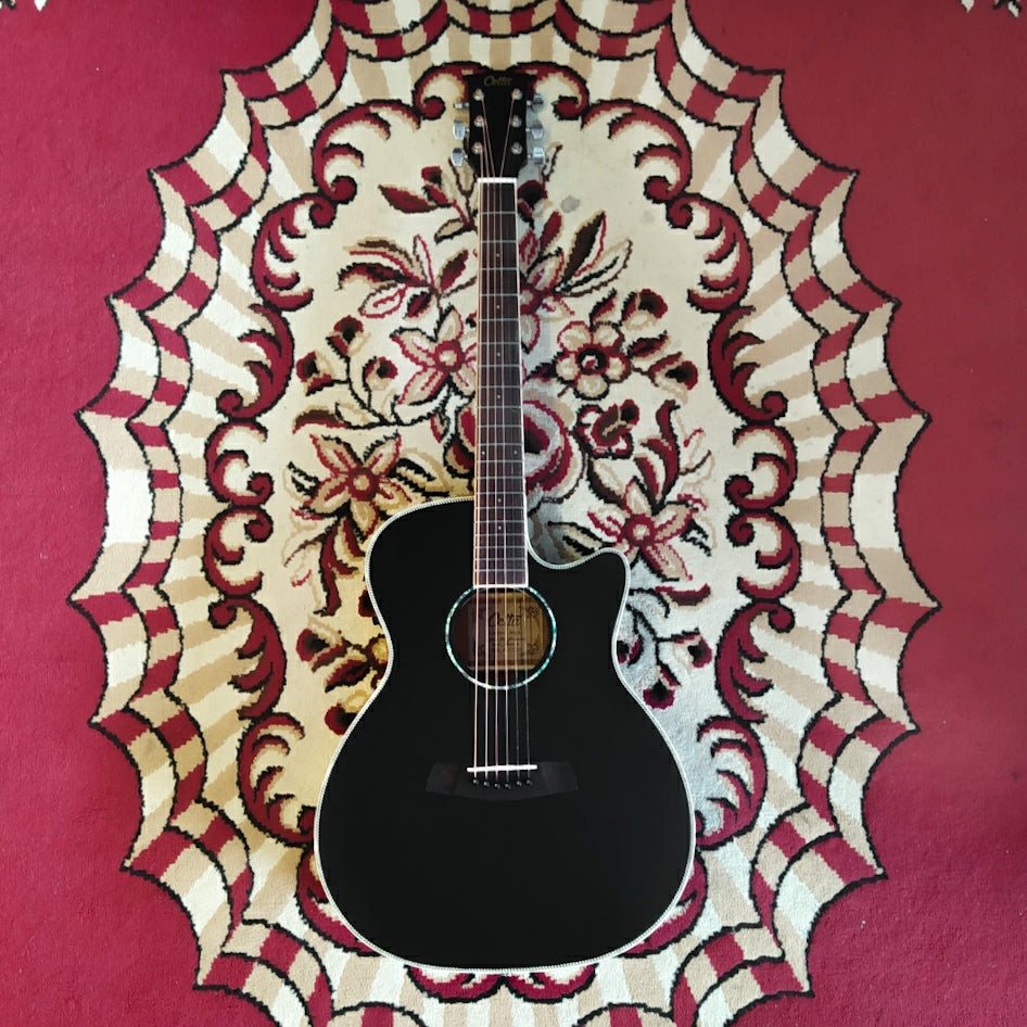 A Cetta OM28CE acoustic guitar resting on a red carpet, featuring a black wood finish and cutaway design.