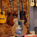 A handcrafted acoustic guitar with barong and rangda motifs on the body, displayed among other guitars against a wooden wall.