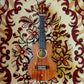 An electric ukulele with a solid acacia body and rosewood fingerboard, placed on a floral-patterned carpet.